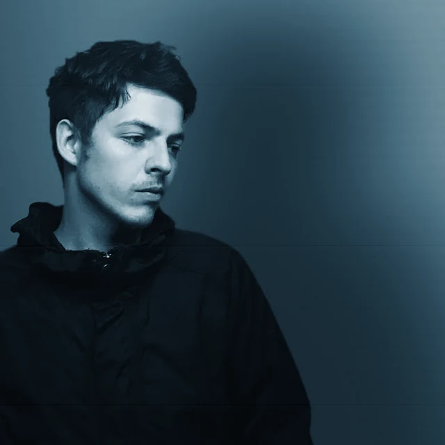 DYL in a dark jacket beside a softly shadowed wall. Photography: Roberta Rotariu. Blue film print treatment.