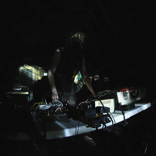 MITH-XX performing with hands over electronic instruments and cables in a dark room with subtle coloured reflections. Archival print treatment.