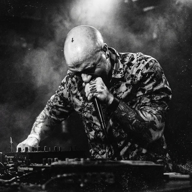 Celldöd performing with a microphone, one tattooed hand holding it and the other operating electronic equipment. Archival print treatment.
