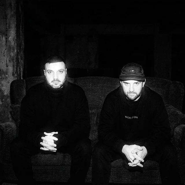 NX1 — Surit and Samot seated together in dark clothing, with their hands clasped. Archival print treatment.