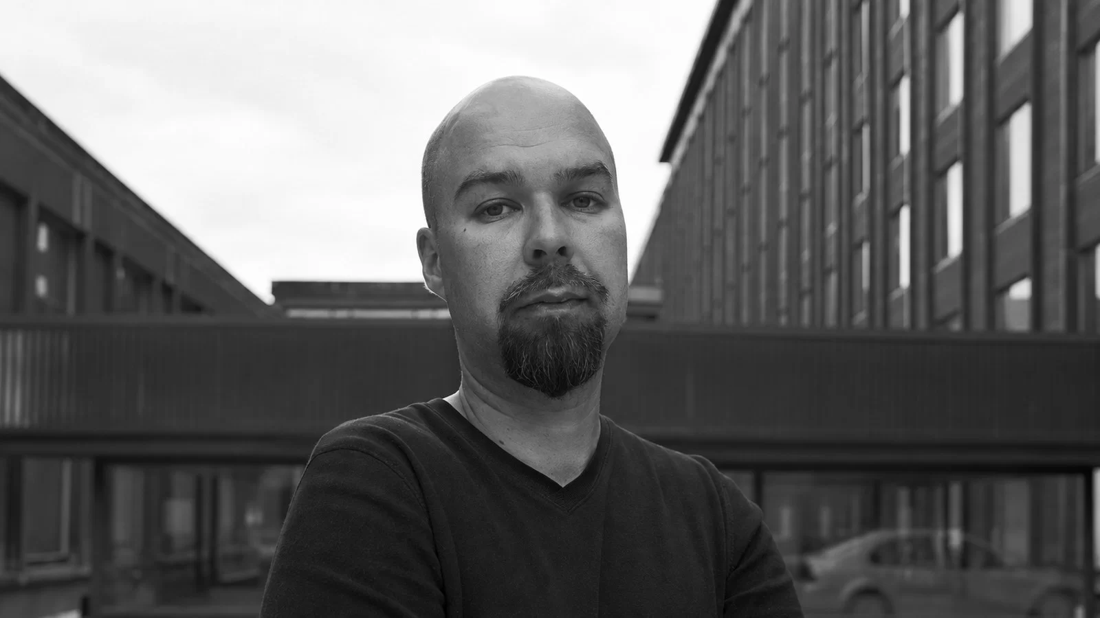 Mika Vainio in a black-and-white portrait outside industrial buildings.