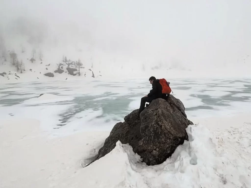 DYL, wearing an orange backpack, sitting on a rock above a snowy lakeside.