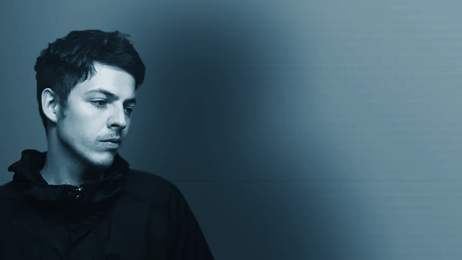 DYL in a dark jacket beside a softly shadowed wall, with a restrained blue horizontal film treatment.