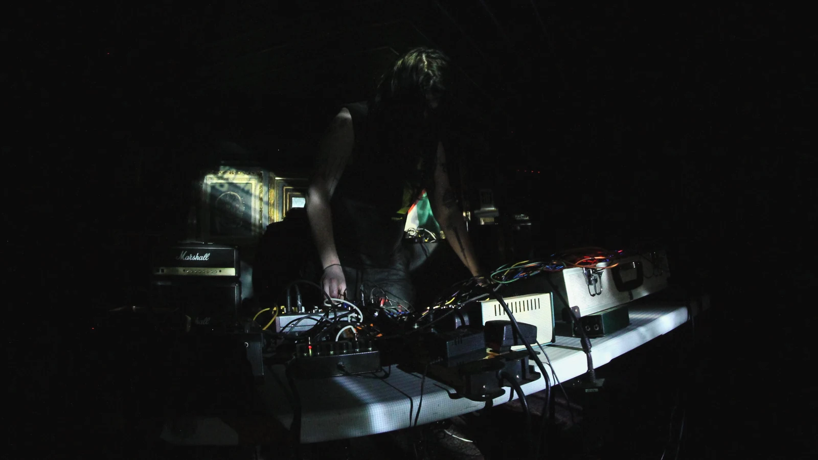 MITH-XX performing with hands over electronic instruments and cables in a dark room with subtle coloured reflections.