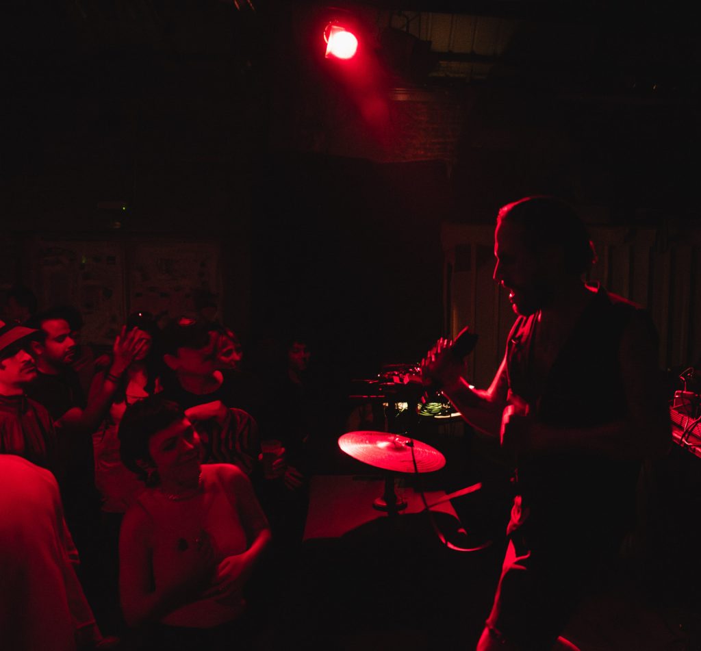 OTTOMAN GRÜW performing beside an audience in a dark room with red lighting.