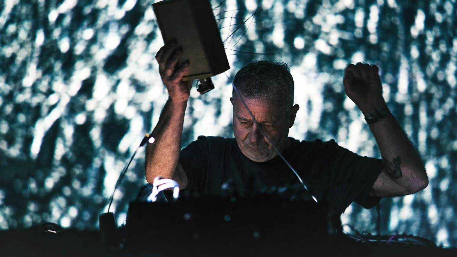 Drew McDowall performing live with raised arms in front of projected light.