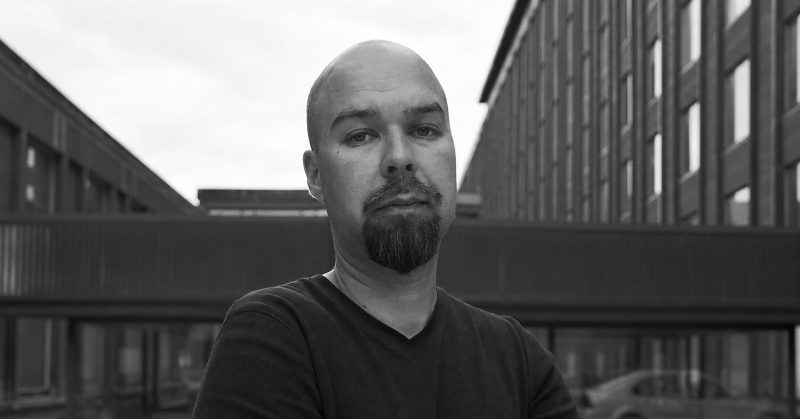 Black-and-white portrait of Mika Vainio outside industrial buildings