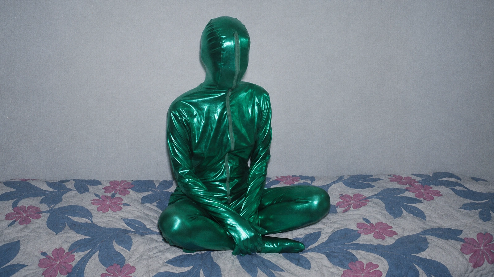 Èlg in an enclosed metallic green suit, seated cross-legged on a muted floral bed against a bare cool-grey wall.