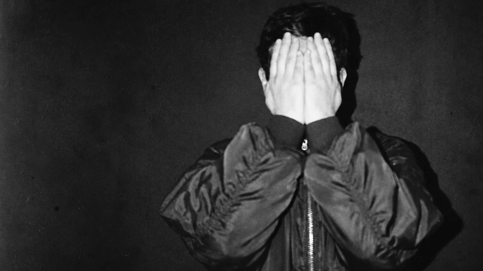 ANFS covering his face with both hands against a dark background.