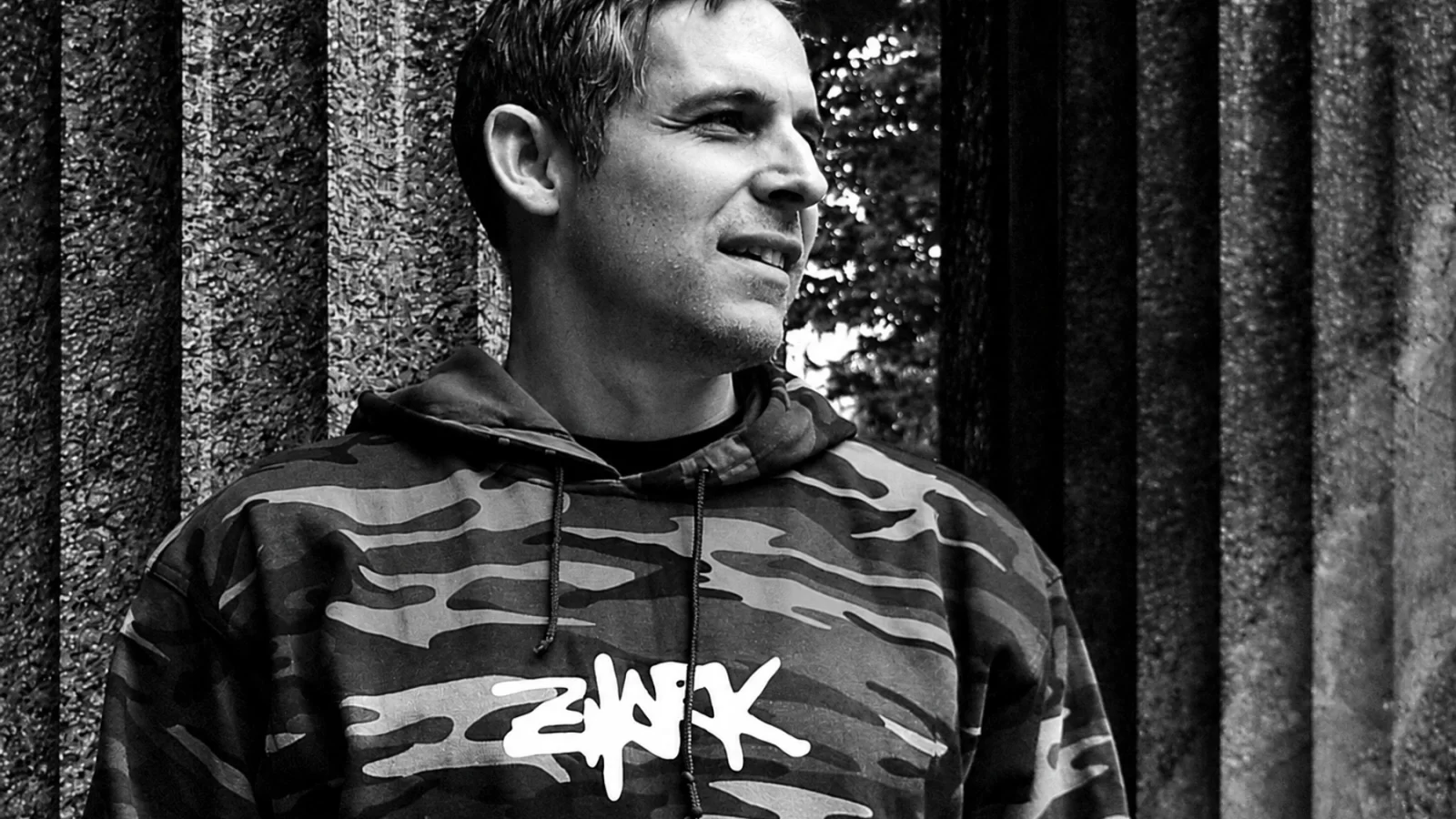 Kareem wearing a Zhark camouflage hoodie beside a concrete wall.