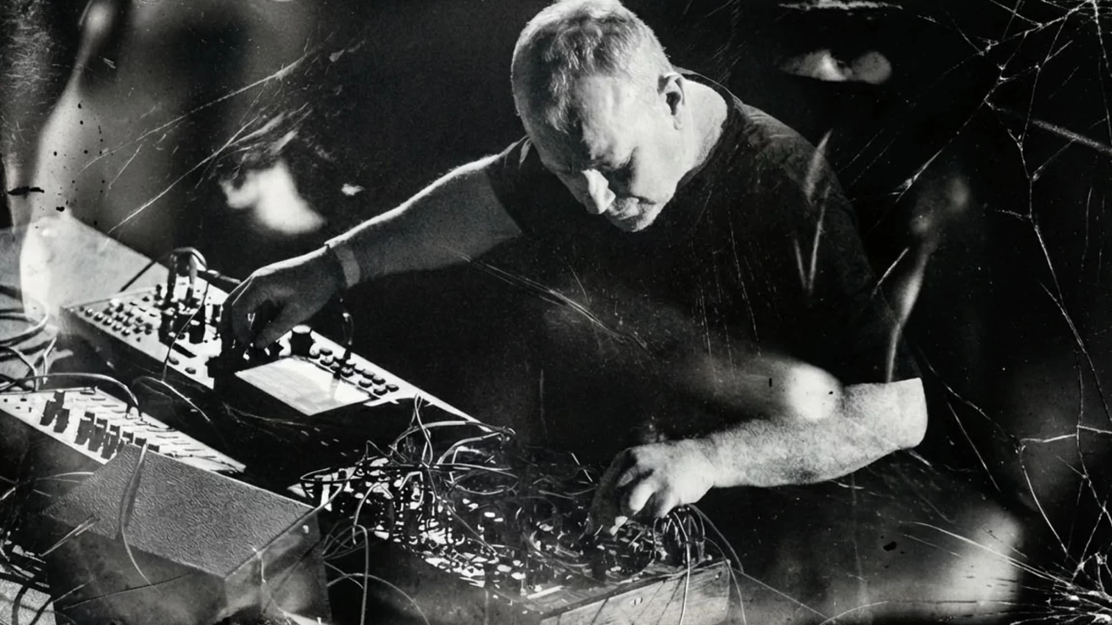 Drew McDowall performing with electronic equipment in a layered black-and-white photograph.