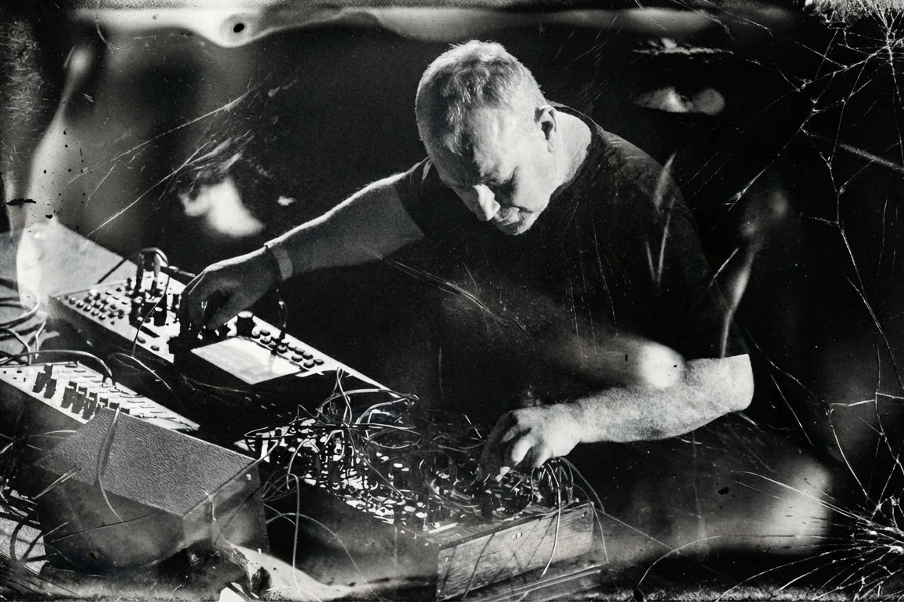 Drew McDowall performing with electronic equipment in a layered black-and-white photograph.