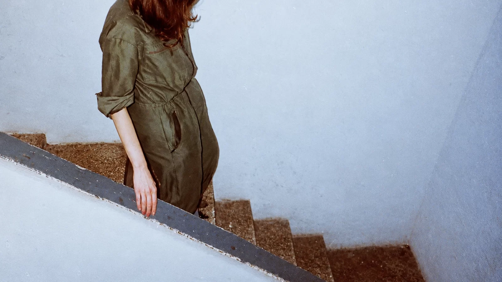 Okkre in an olive jumpsuit on a stairway, photographed against a pale blue wall.