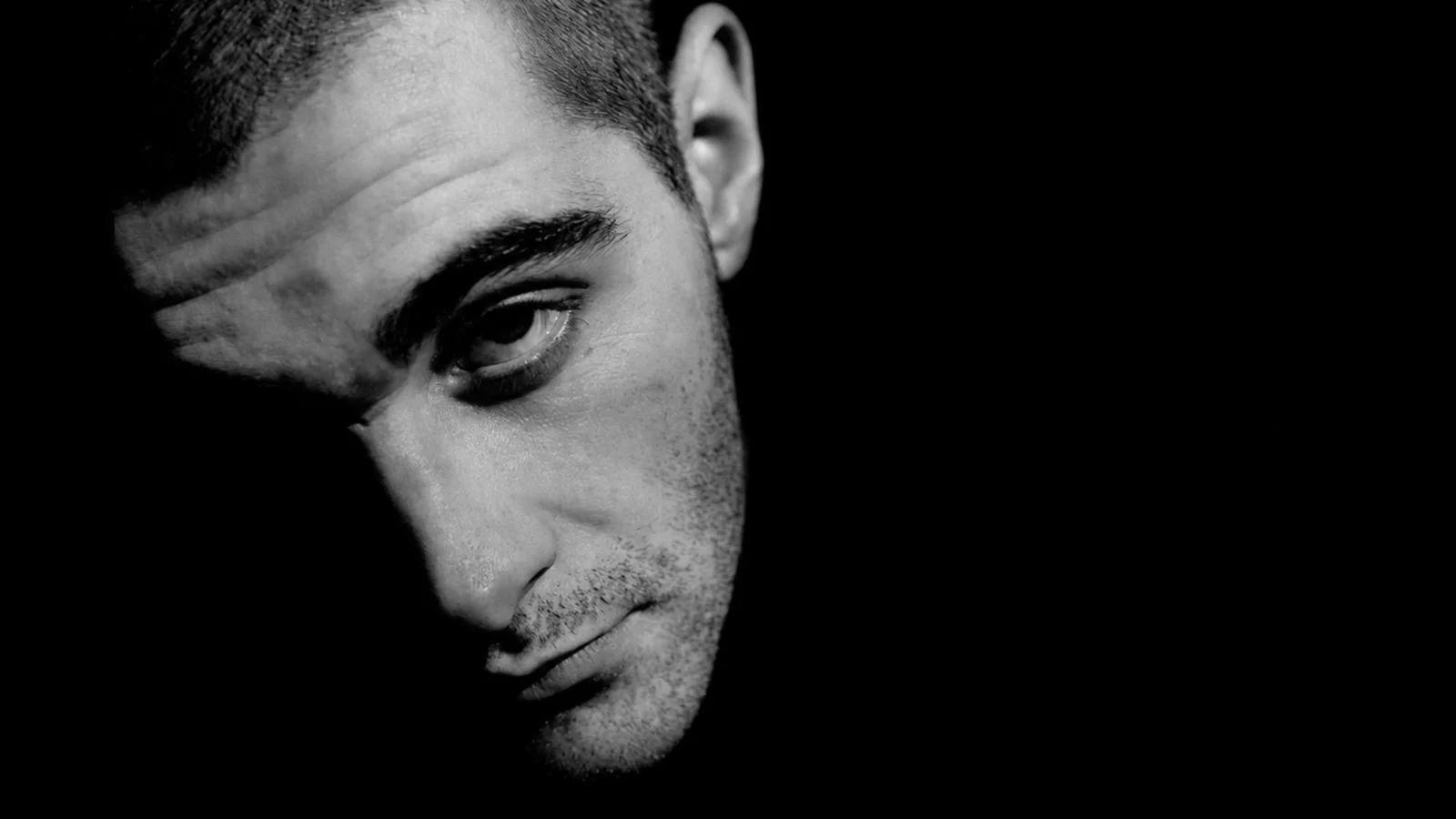 Paulie Jan’s face emerging diagonally from deep shadow in a monochrome close-up.