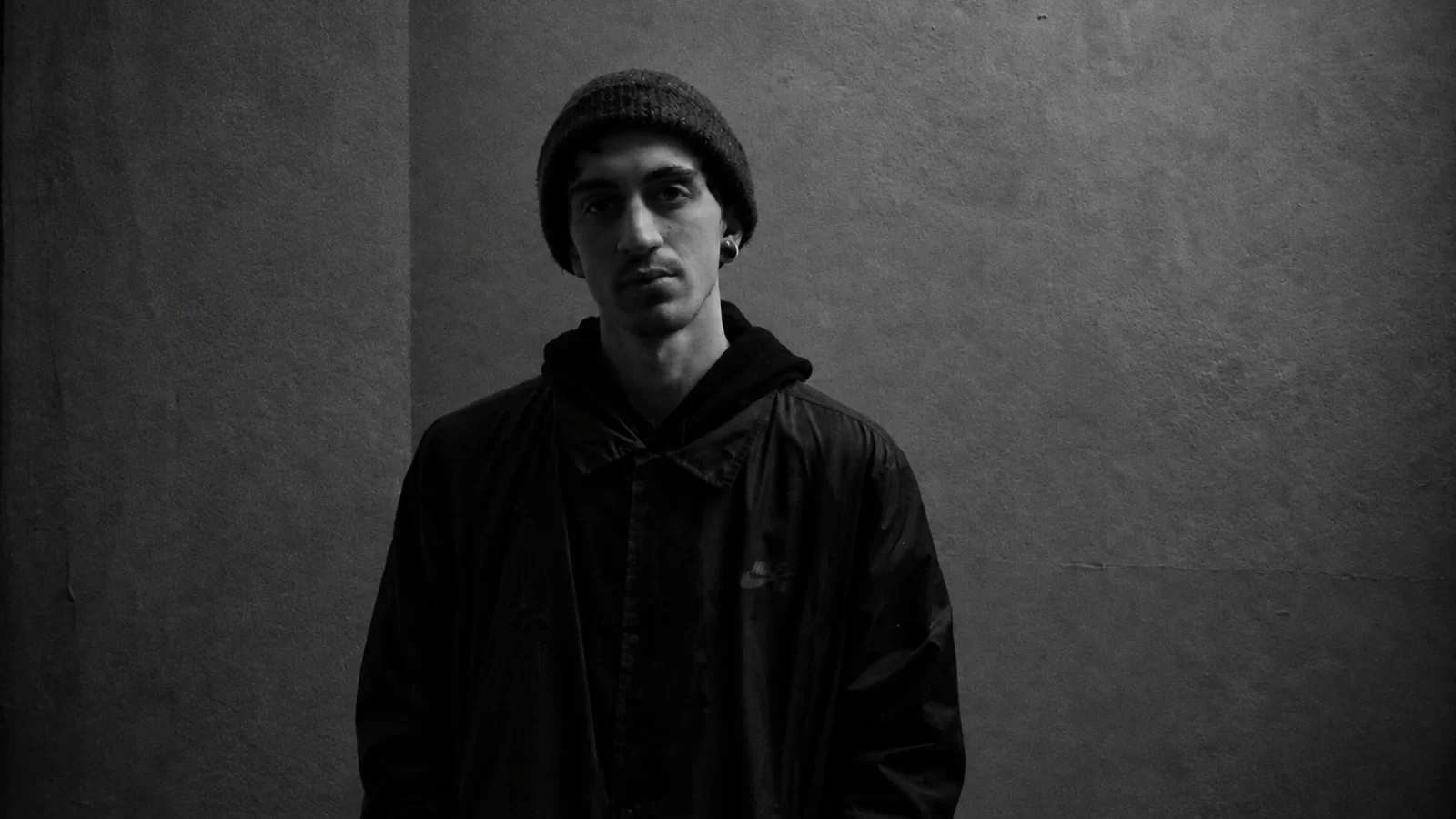 Giulio Spampinato / Coeden in a black beanie and jacket against a textured wall.
