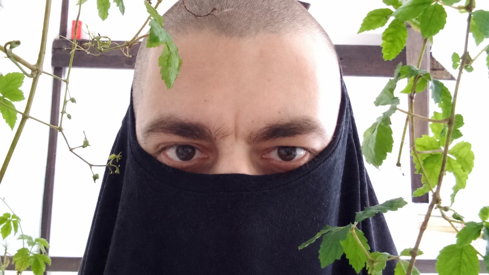 N1L with his face partly covered by dark fabric, framed by green plants.