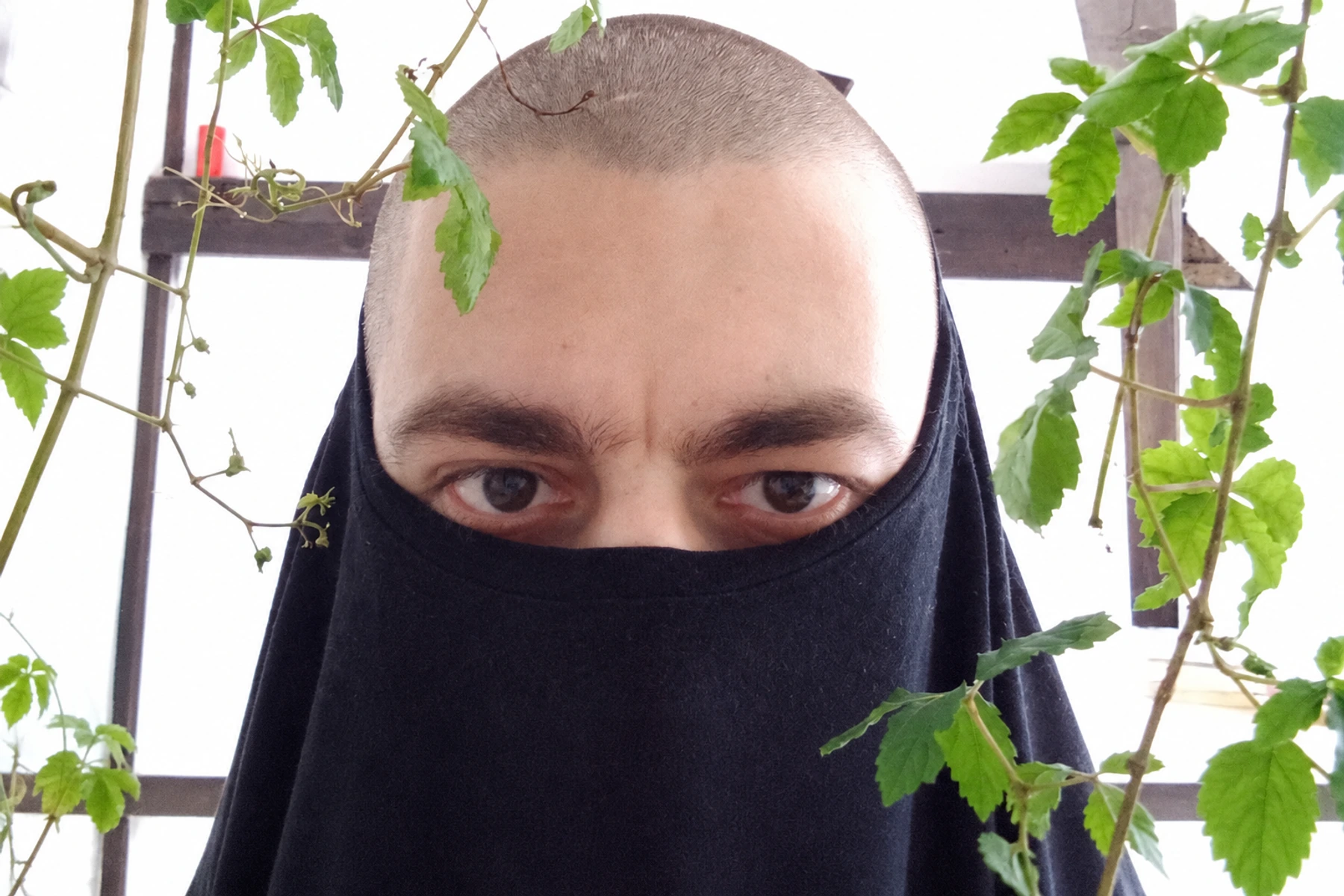 N1L with his face partly covered by dark fabric, framed by green plants.