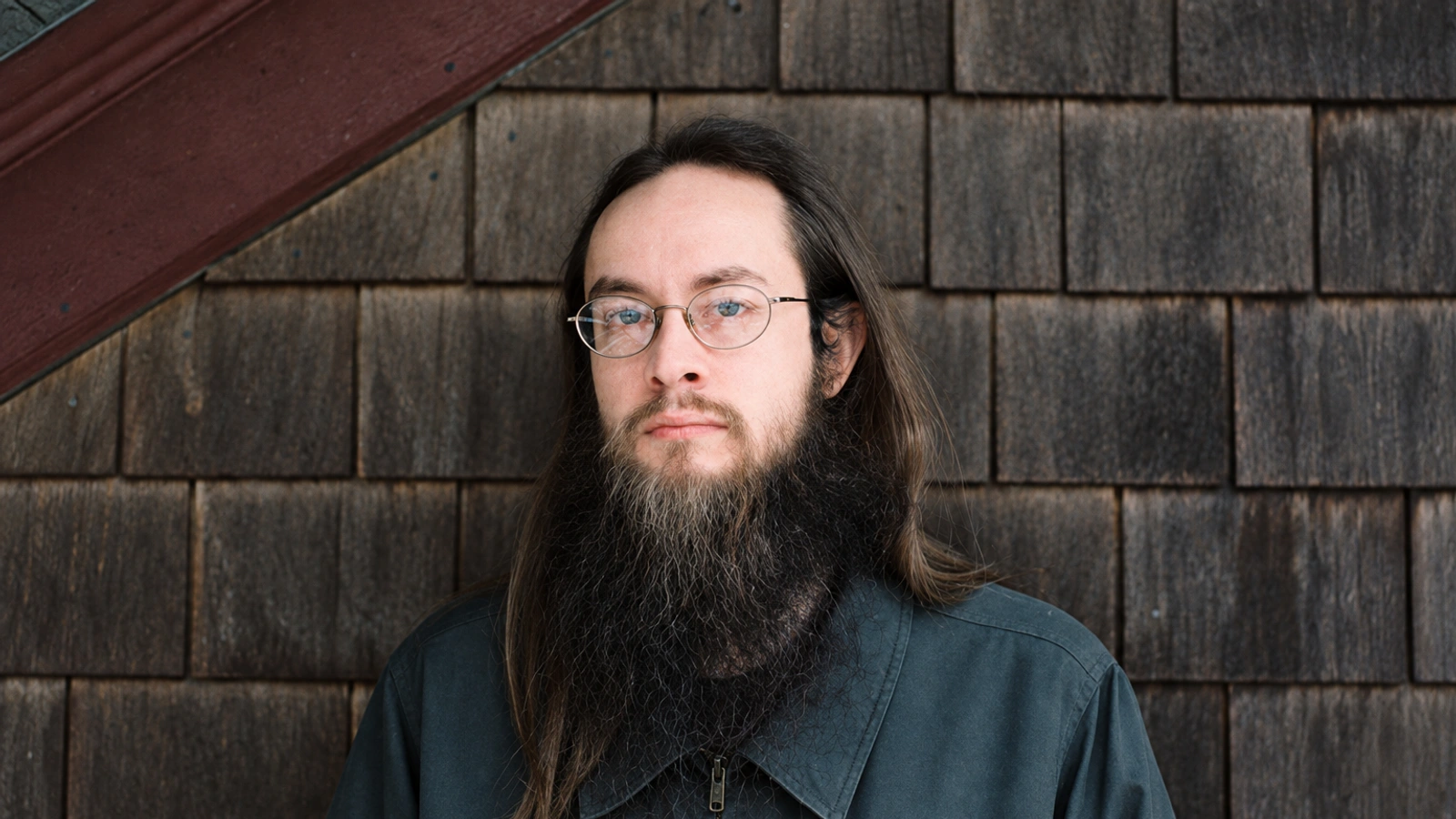 Aaron Martin with long hair, beard and glasses in a cool-toned portrait against weathered shingles.
