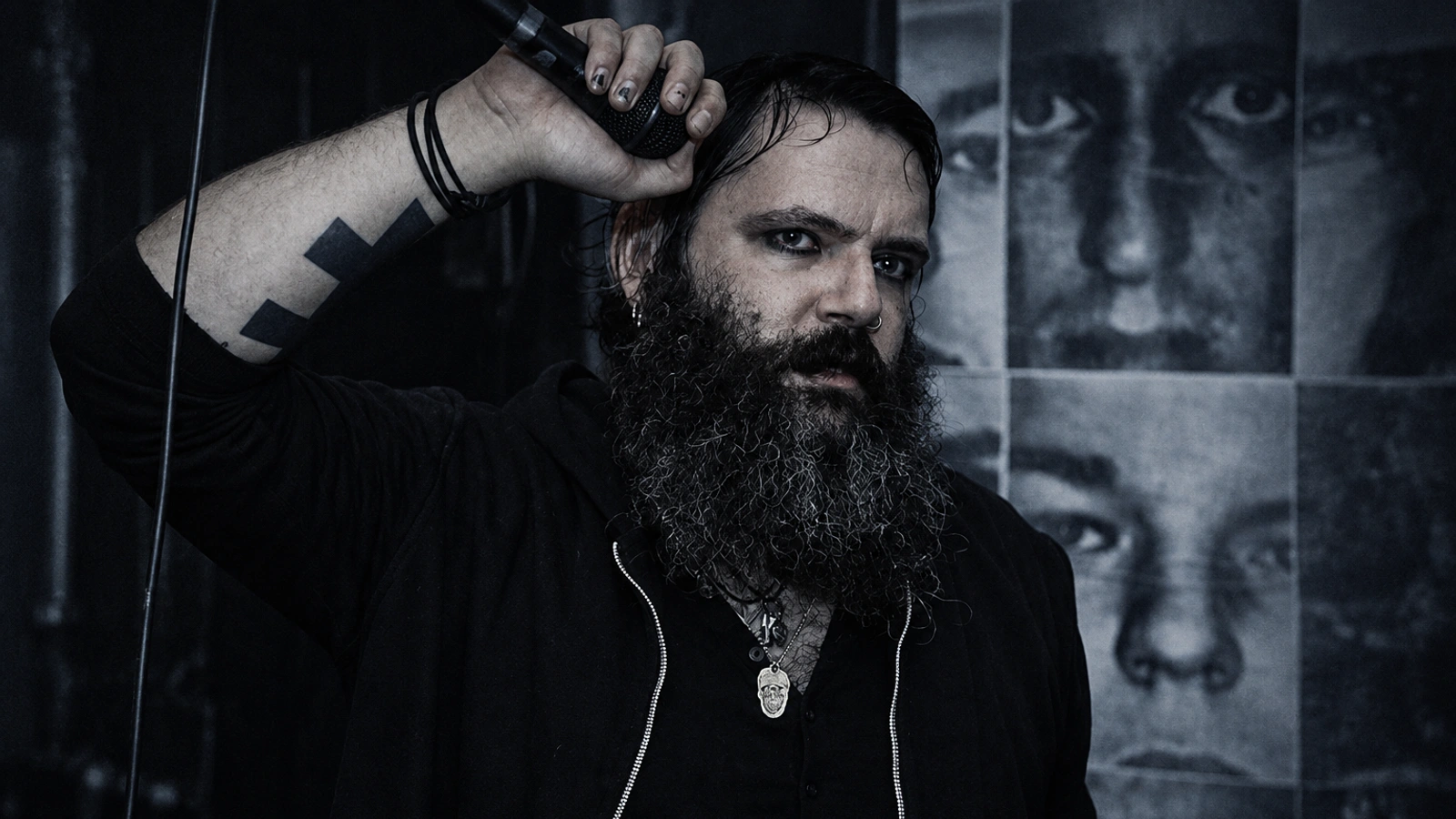 Thomas Martin Ekelund with long beard, raised tattooed arm and black hoodie against a wall of portraits.