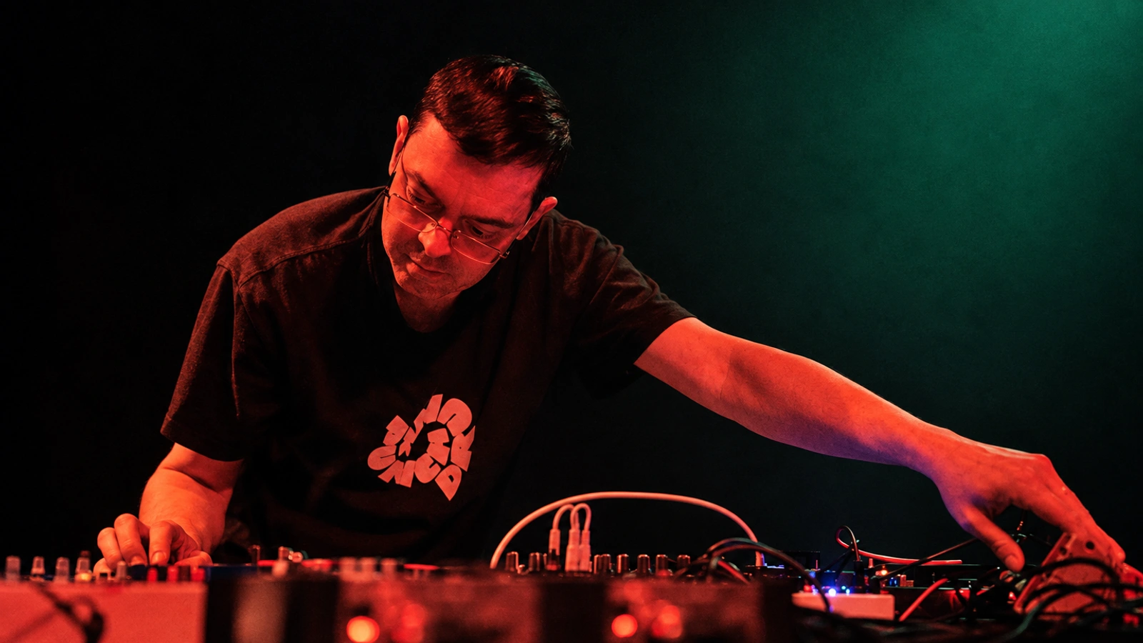 Ekoplekz working at electronic instruments under red light and a dark teal stage glow.