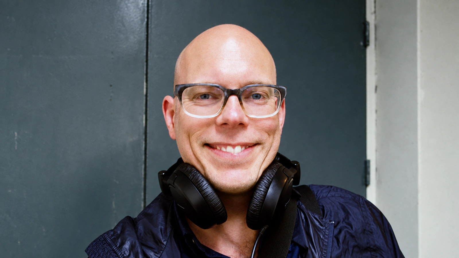 BJ Nilsen wearing glasses and headphones around his neck against a cool grey door.