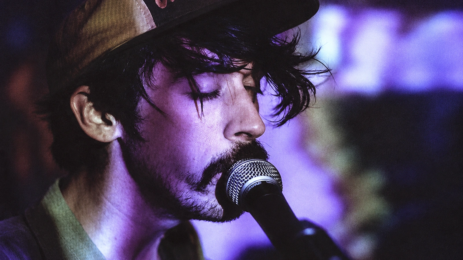 Sun Araw singing into a microphone beneath deep violet stage lighting.