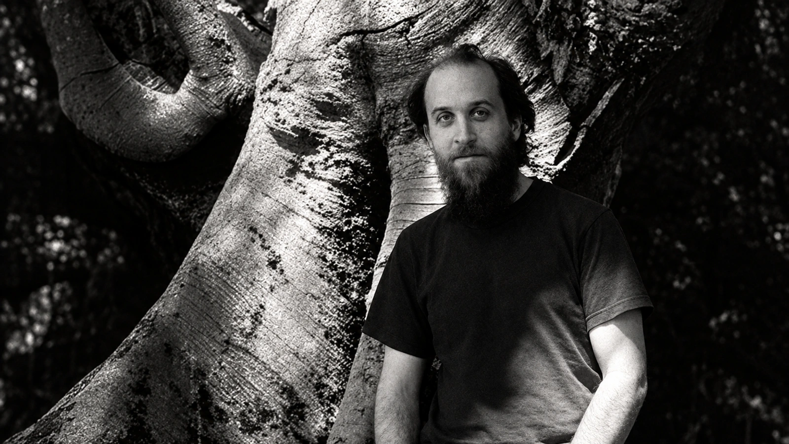 Aidan Baker standing beside a tree in a grainy black-and-white portrait.