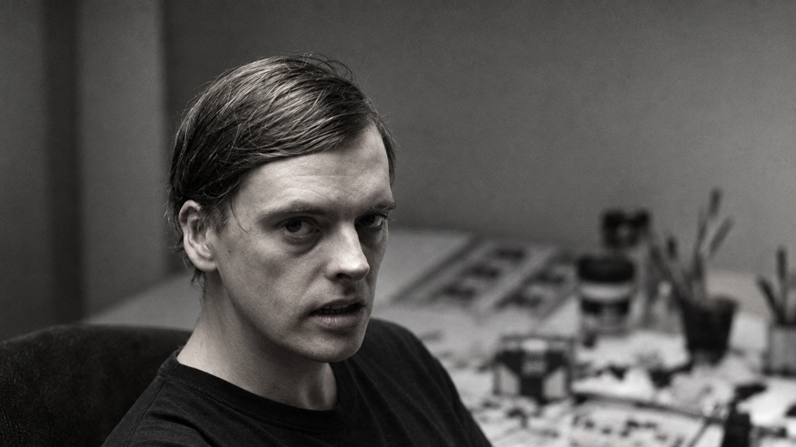Mark Harwood seated at a creative worktable, looking toward the camera in a monochrome portrait.