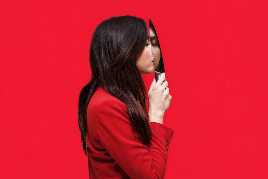 Camella Lobo in a red jacket holding a narrow mirror against a red background.