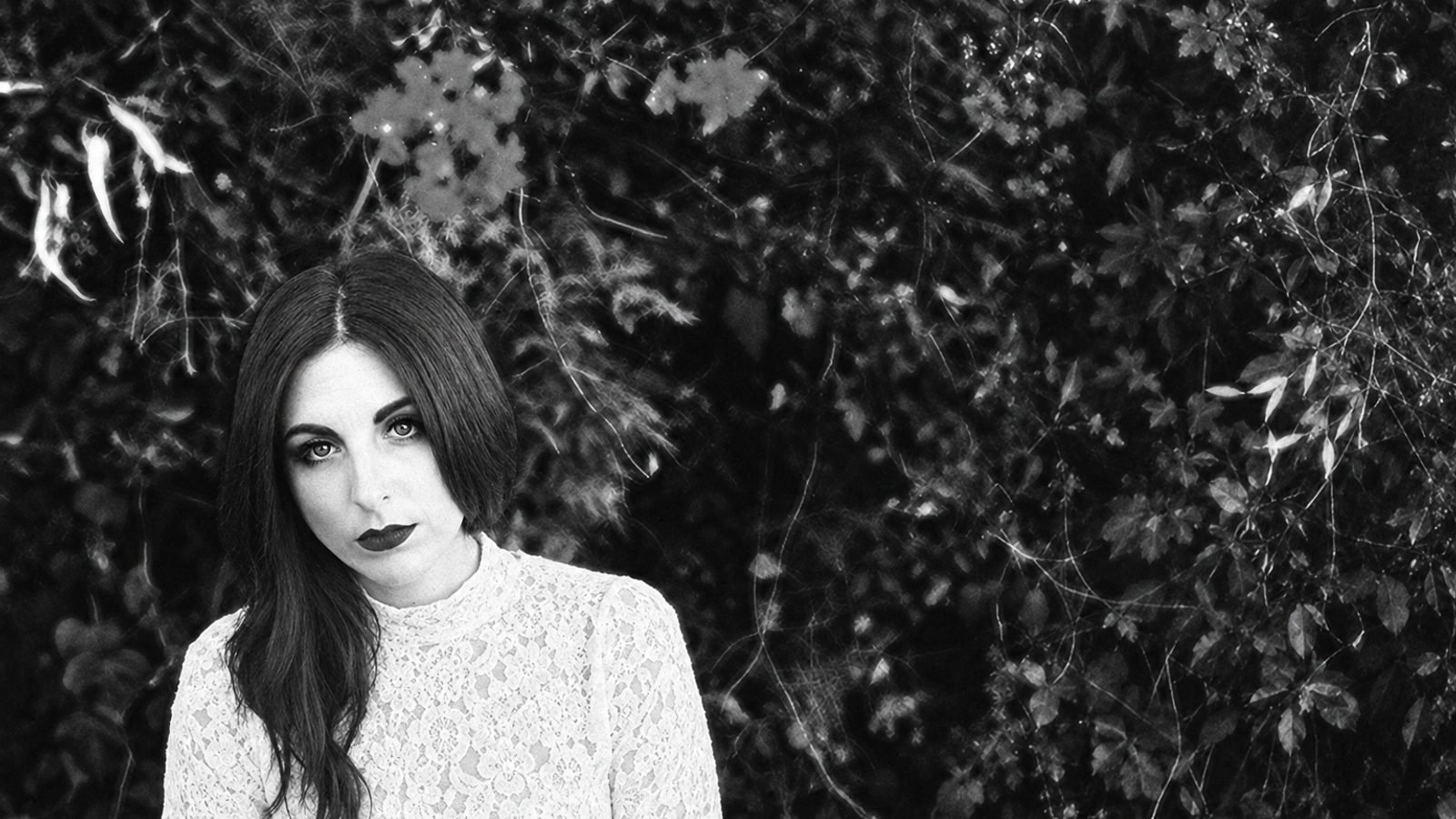Camella Lobo of Tropic Of Cancer in a white lace top beneath dark foliage.