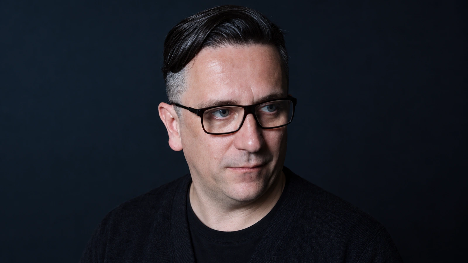 Peter Rehberg wearing black-framed glasses, a black crew-neck shirt and a dark cardigan against a muted blue-black backdrop.