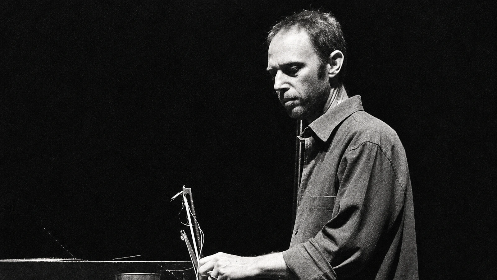 Steve Roden performing with small objects on a dark stage, in a grainy monochrome photograph.