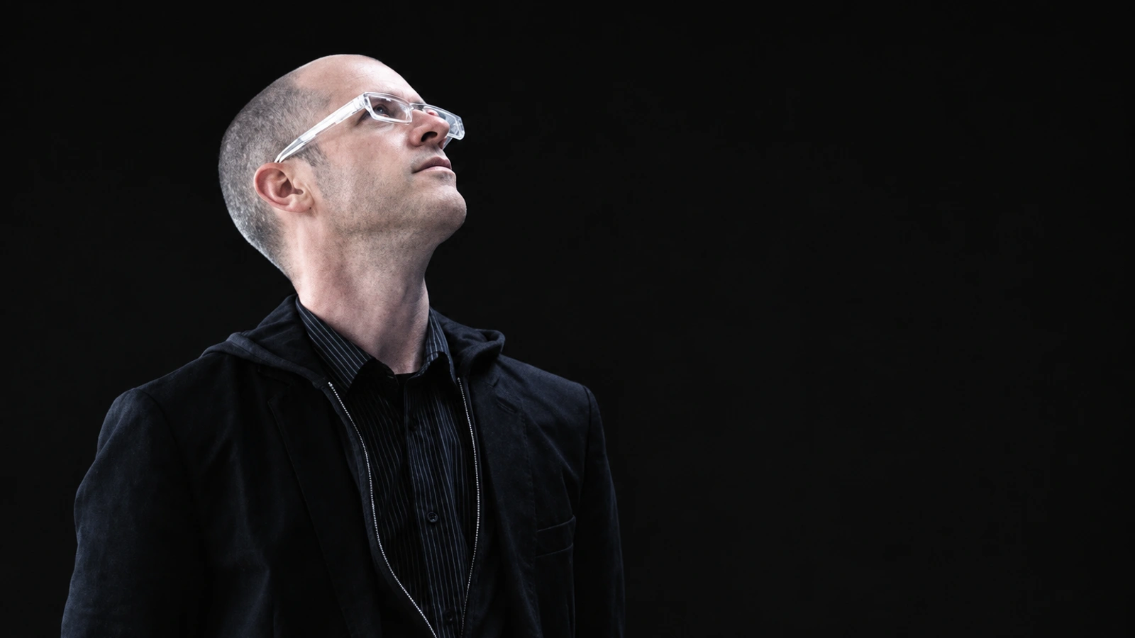 Richard Chartier looking upward through clear glasses against a deep black background, with restrained cool tones.