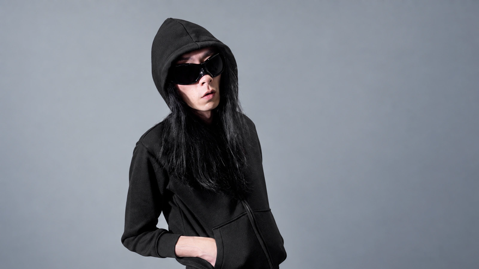 Xhin in a black hood and sunglasses against a cool grey background.