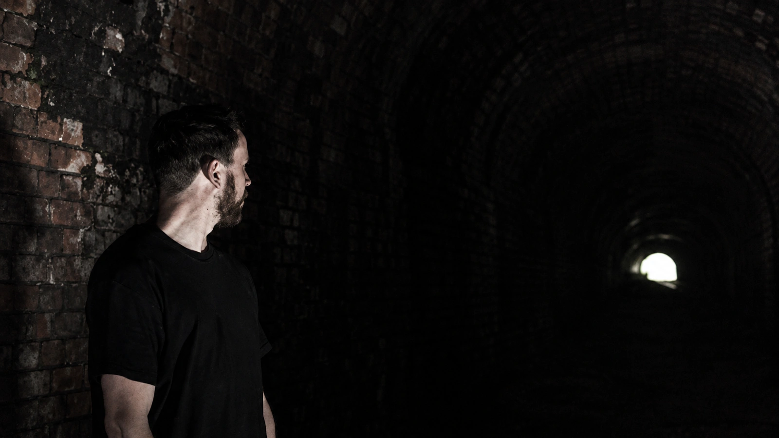 Roly Porter standing inside a dark railway tunnel, with a small bright opening in the distance.