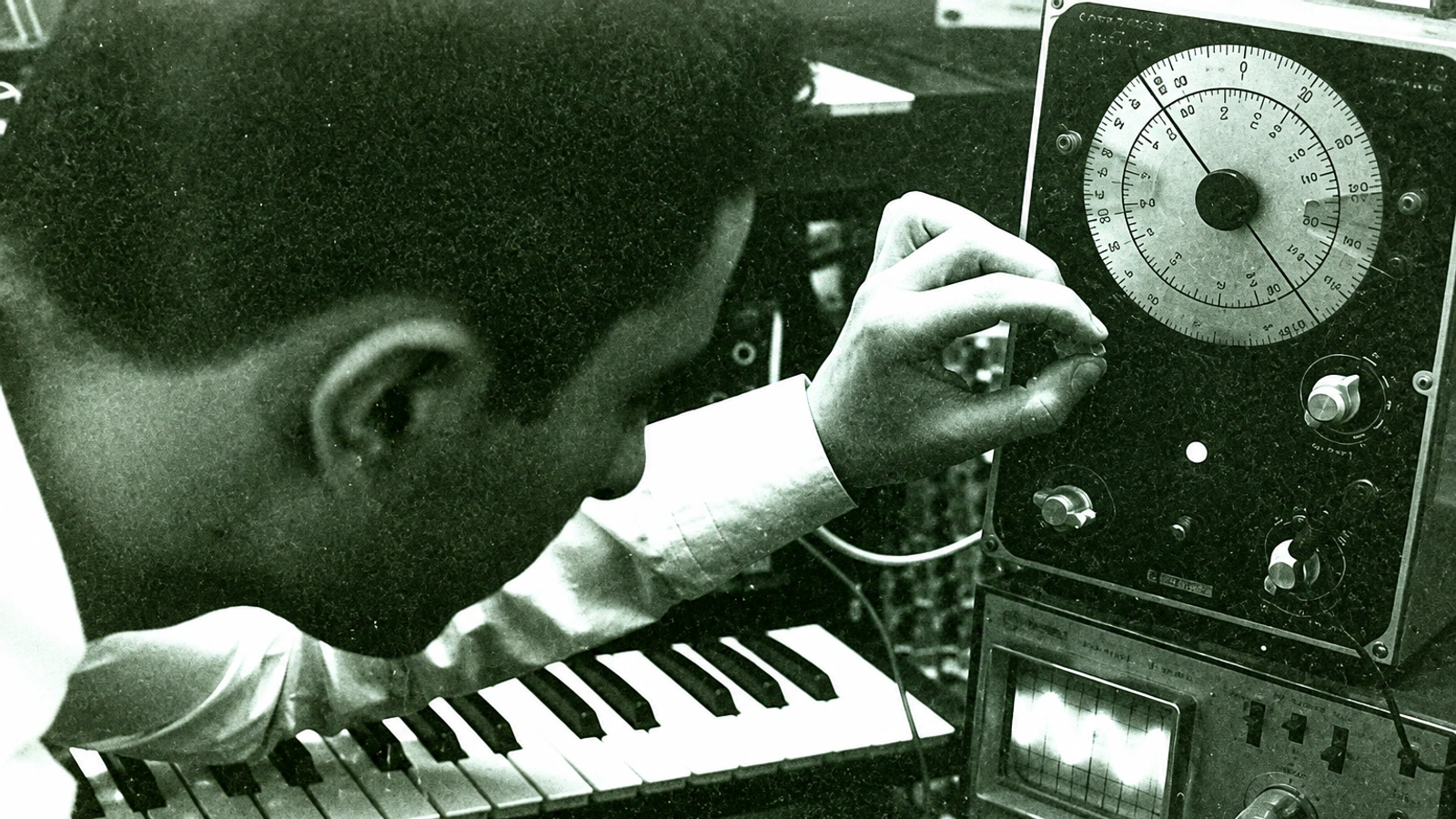 Geistform adjusting an analogue oscillator beside a keyboard in a green-tinted archival photograph.