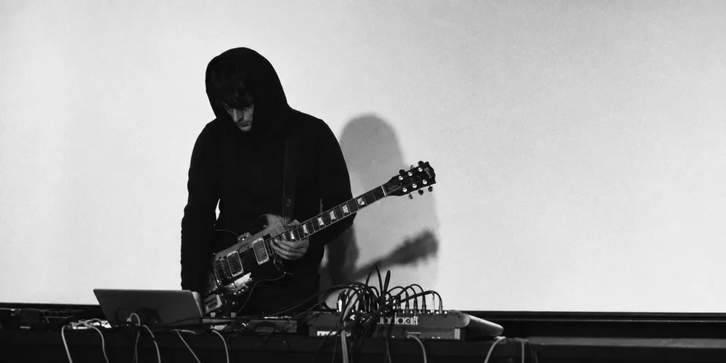 The Sight Below playing guitar behind tabletop electronics, his shadow on the wall.