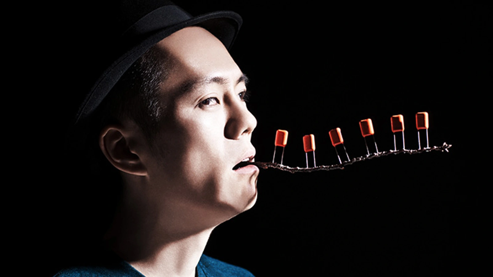Sugai Ken in profile holding a small line of electronic components in his mouth.