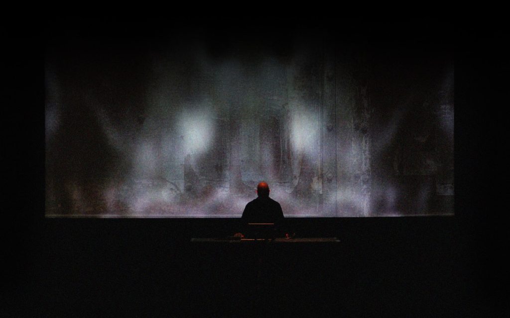 Lustmord performing in silhouette before a large atmospheric projection.