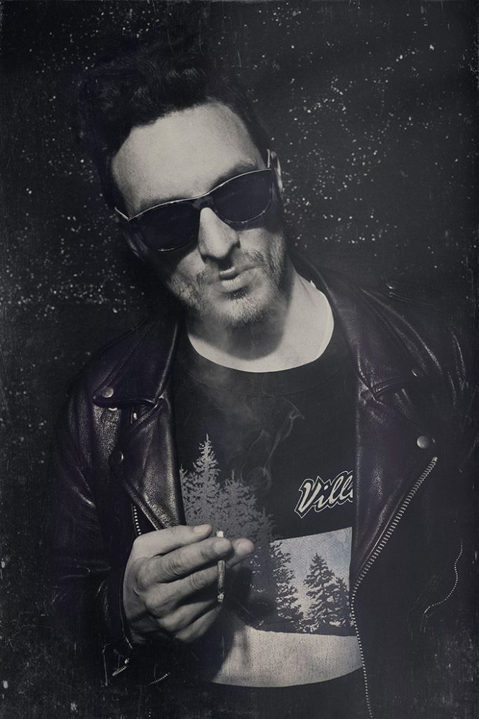 Ernestas Sadau wearing sunglasses and a leather jacket, holding a cigarette in a vintage portrait.