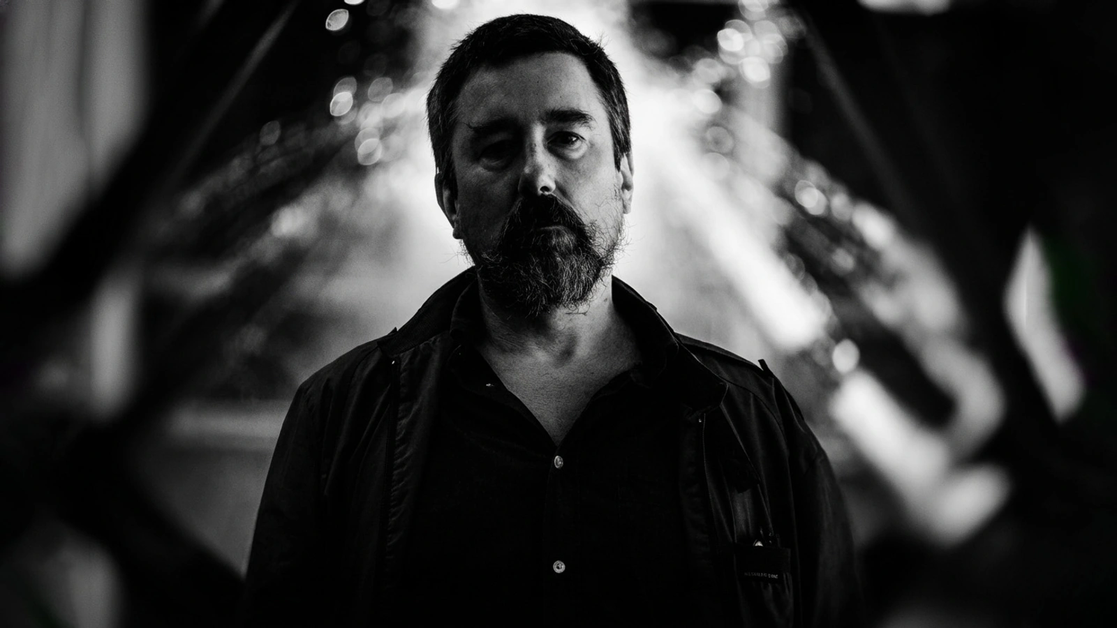 Black To Comm in a monochrome portrait with blurred architectural forms behind him.