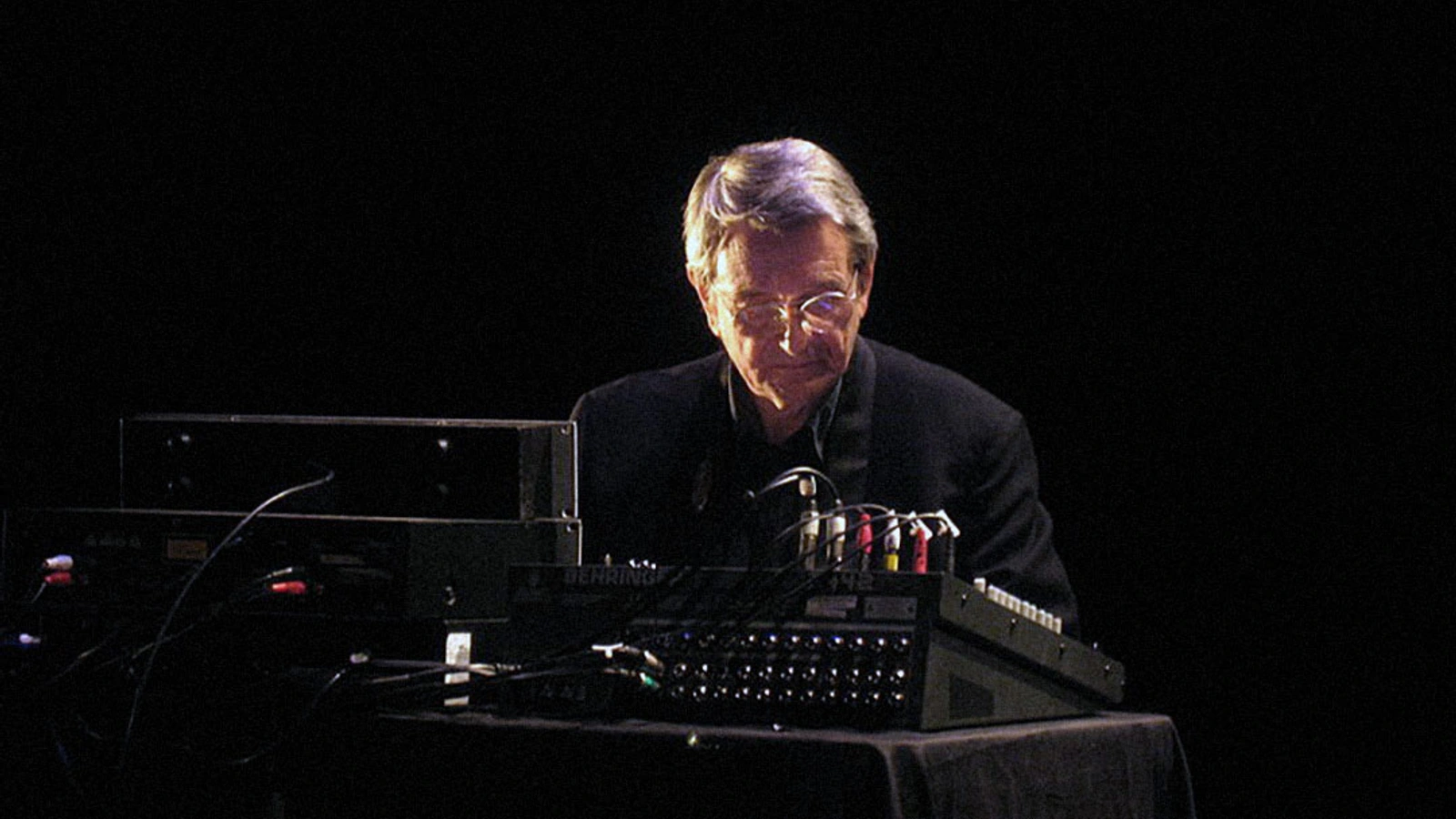 Asmus Tietchens performing behind electronic instruments on a dark stage.