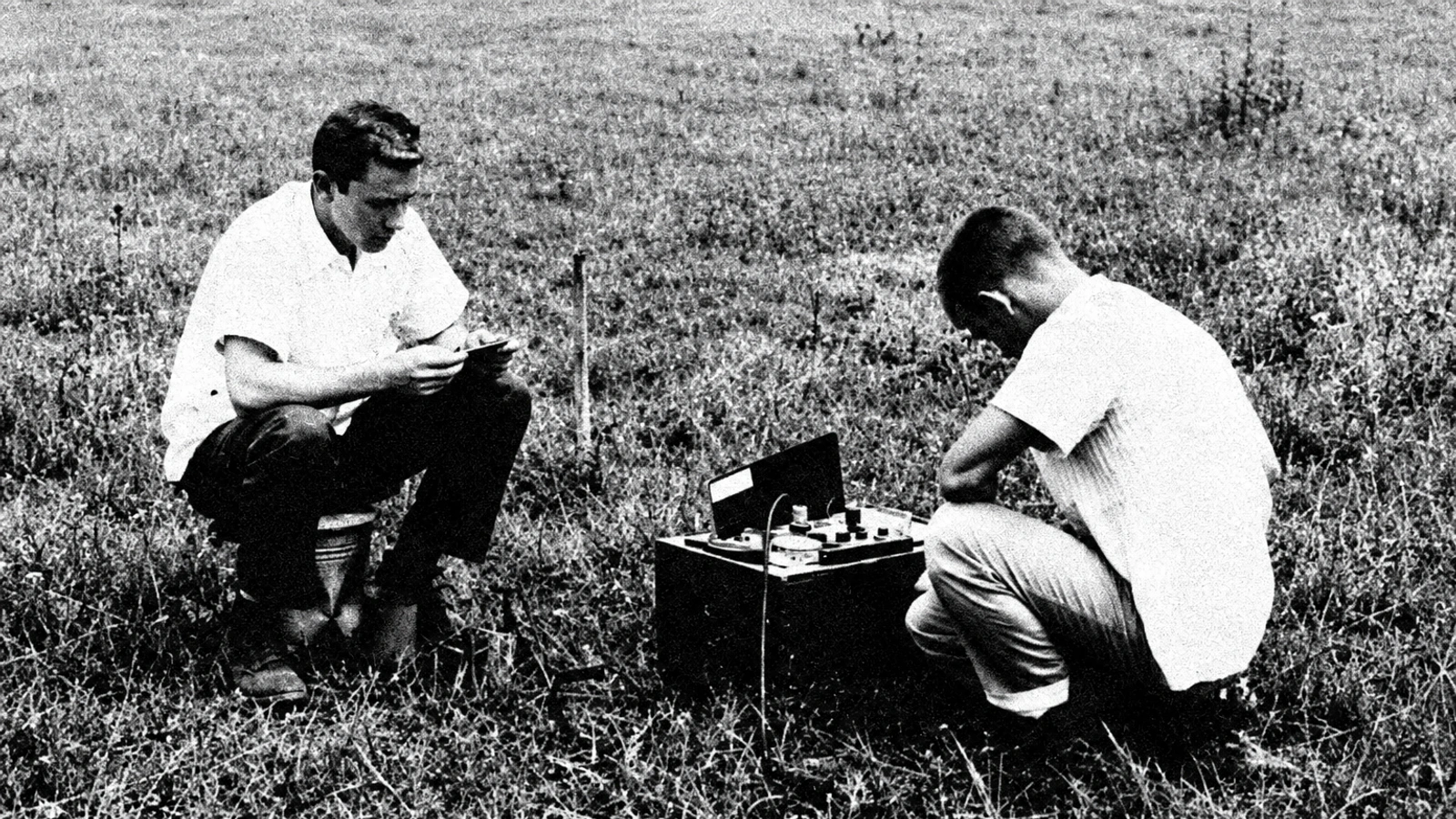 Two figures with a record player in an open landscape, in a grainy archival photograph.