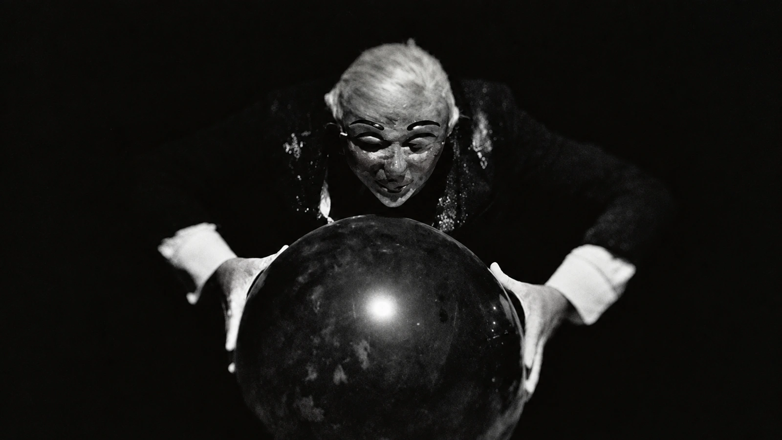 Kreng theatrical portrait with a pale mask, wild white hair and hands around a dark sphere.