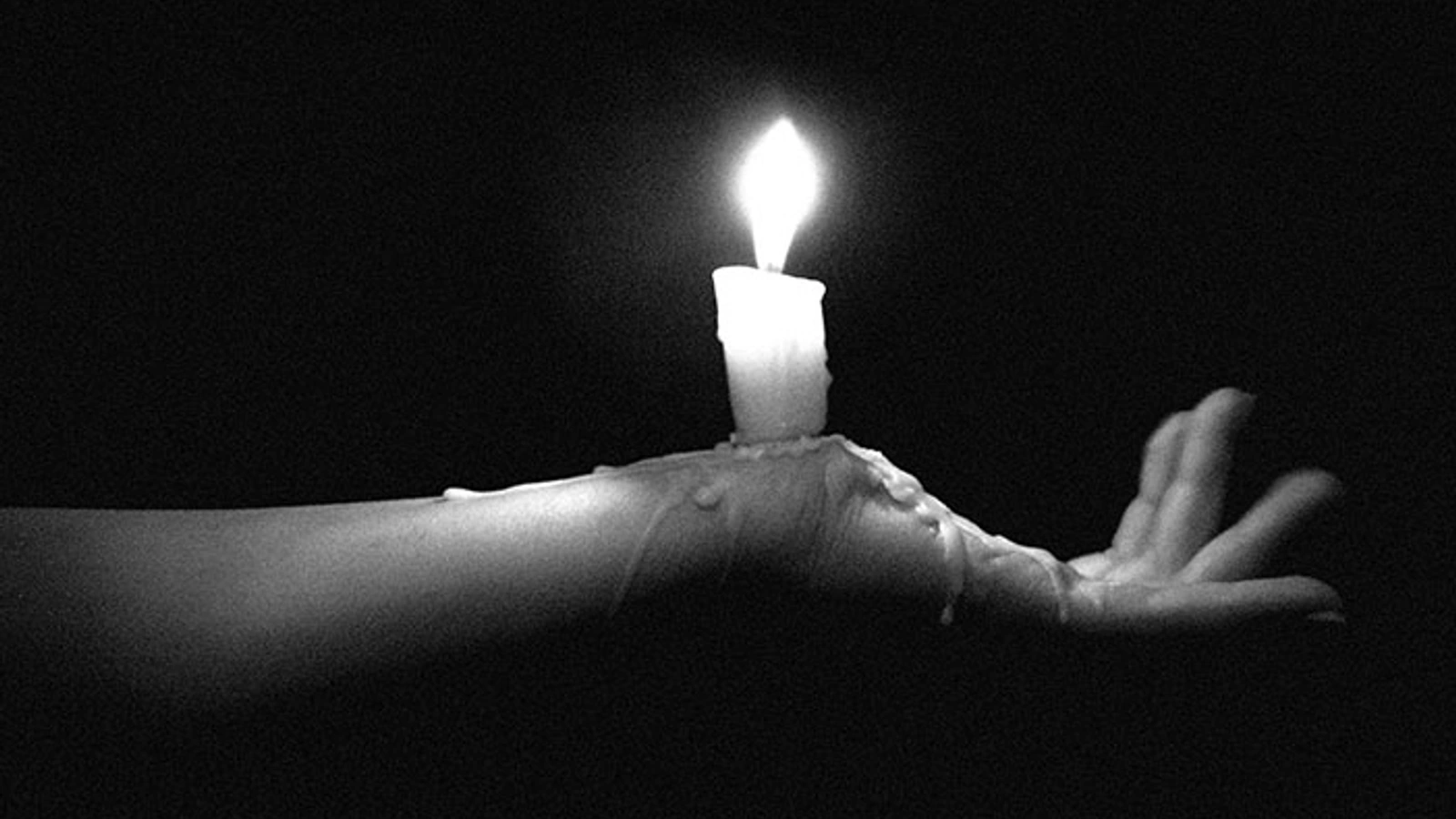 A burning candle held in a hand covered in dripping wax, photographed in grainy black and white.
