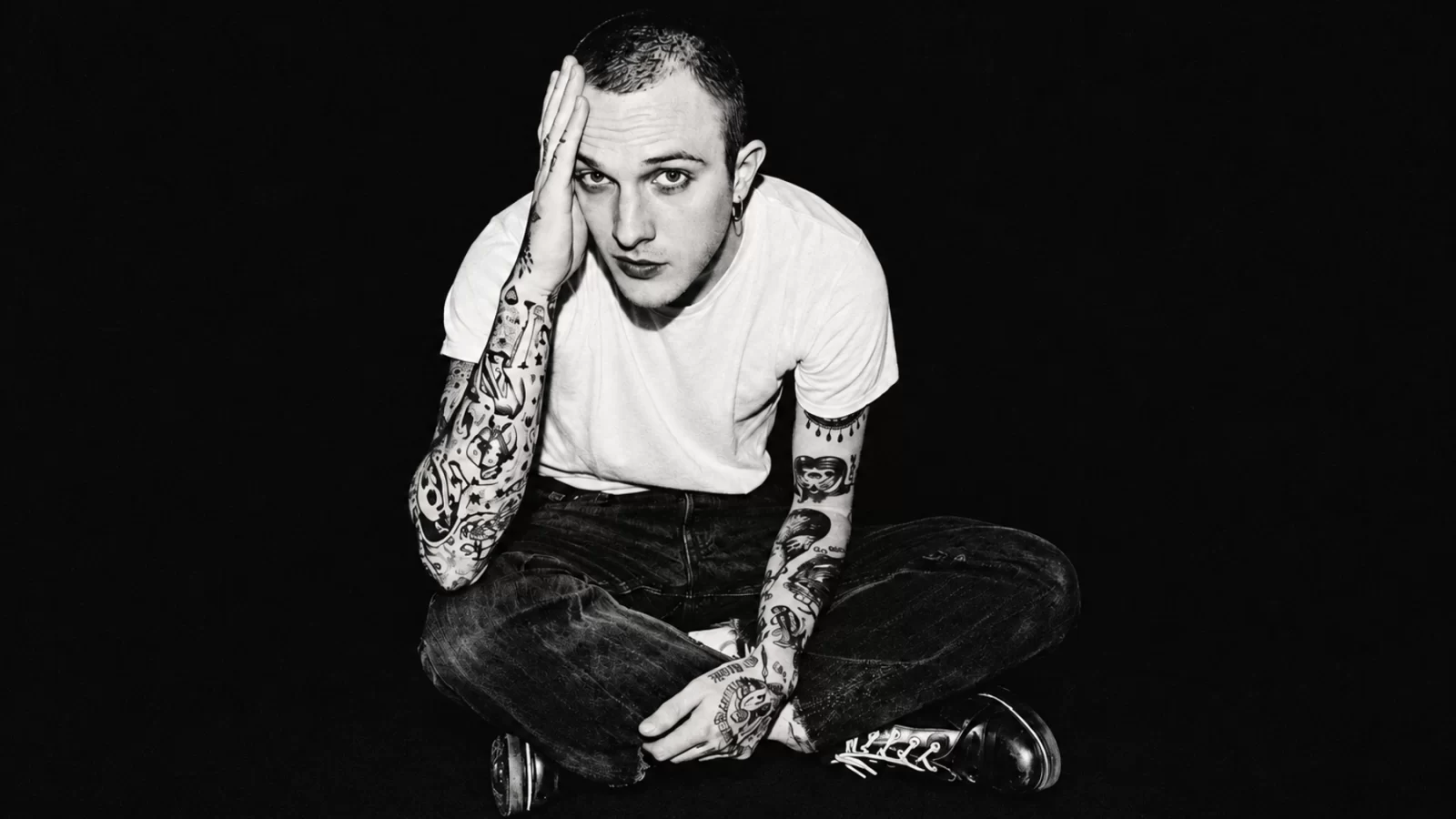 M!R!M sitting cross-legged in a white T-shirt, one hand against his face, against a deep black background.