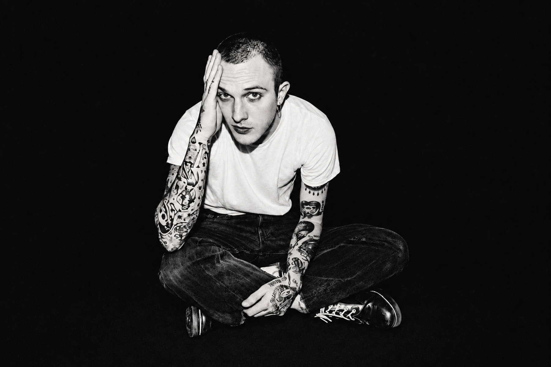 M!R!M sitting cross-legged in a white T-shirt, one hand against his face, against a deep black background.