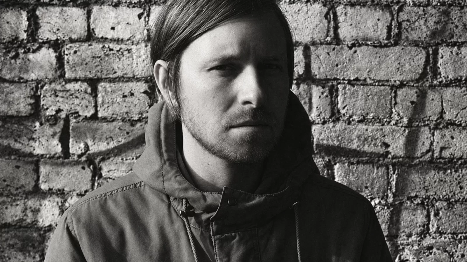 Black-and-white portrait of Blanck Mass in a dark coat against a brick wall.