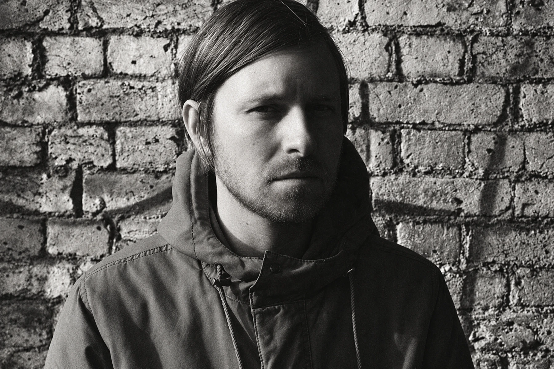 Black-and-white portrait of Blanck Mass in a dark coat against a brick wall.