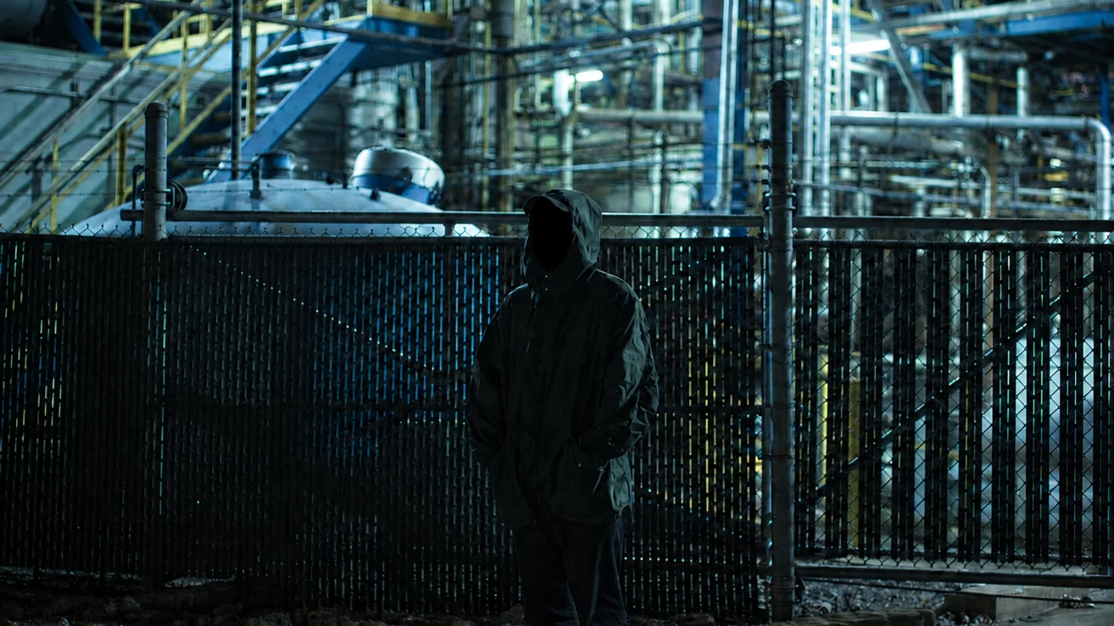 LXV with his face completely in shadow beside an illuminated industrial structure.